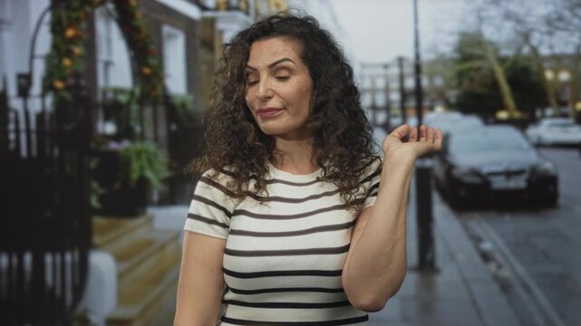 Woman wearing striped top pulls earlobe on urban street with parked cars and wrought iron railings; confidence.
