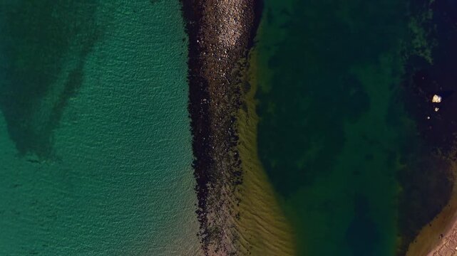 Waves gently lap against the sandy beach as vibrant turquoise waters meet deeper greens. Shadows dance along the rocky edge under the bright afternoon sun, creating stunning contrast.