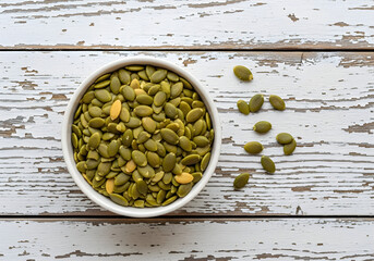 Top view of organic green pumpkin seeds in a white bowl on a rustic wooden background