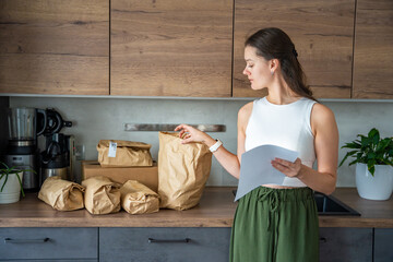 Young woman checking a list while looking at unpacked meal kit ingredients organized by recipe. Concept of planning weekly cooking using convenient pre-portioned grocery delivery.