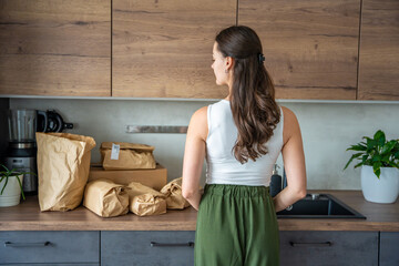 Back view of young woman standing in kitchen and looking at several paper bags with food delivery meal kits. Sustainable meal prep subscription concept.