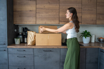 Young woman unpacking a box with pre-portioned ingredients for cooking on the kitchen counter. Concept of meal kit delivery service and easy home cooking with fresh food.
