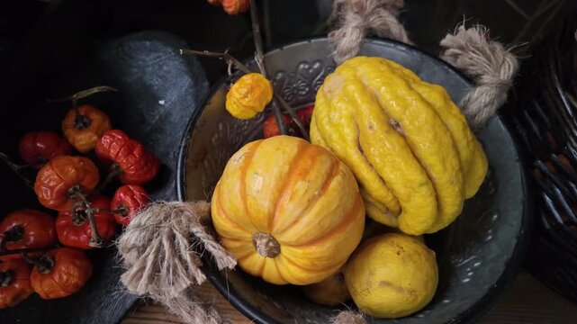 A striped pumpkin, a large yellow citron with protrusions, known as the "Buddha's hand", small red chili peppers lie in a dark metal bowl with decorative elements and coarse twine.