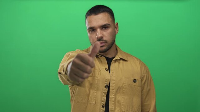 Young hispanic man wearing a mustard jacket displays a thumbs up gesture against a green studio screen; approval.