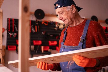 Portrait of Skilled senior elderly carpenter wearing blue coveralls holding wooden plank ready to assemble furniture in small workshop