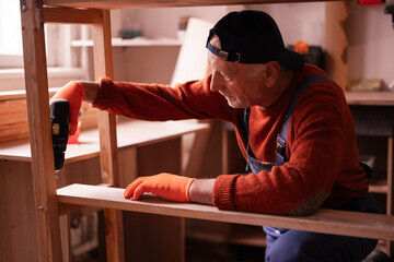 Profession, art and hobby concept. Elderly man carpenter assembling wooden shelf using electric bit...