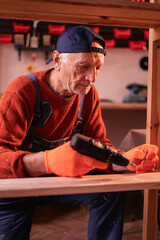 Profession, art and hobby concept. Elderly man carpenter wearing blue overalls assembling wooden shelf using electric drill working on wood in garage. Authentic workshop.