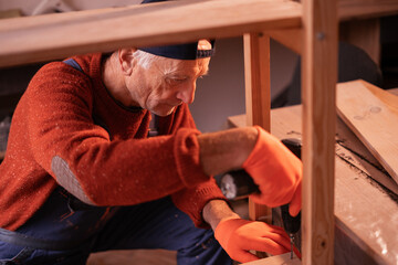 Profession, art and hobby concept. Elderly man carpenter wearing blue overalls assembling wooden shelf using electric drill working on wood in garage. Authentic workshop.
