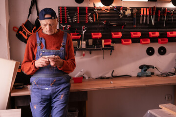 Elderly carpenter chatting online using smartphone in authentic woodworking workshop