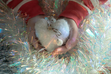 Child hands cradle a fluffy felt heart amid iridescent tinsel; selective focus and soft light tell a cozy lifestyle story and supply heartfelt Christmas elements for happy new year designs. ​