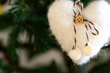 Close‑up heart ornament with wooden button and pom‑poms on evergreen; gentle light, selective focus and soft bokeh create a warm holiday background for Christmas elements and happy new year.