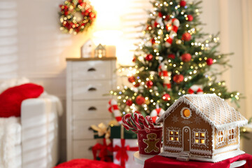 Beautiful gingerbread house and cup with candy canes on table against blurred lights indoors, space for text. Christmas atmosphere