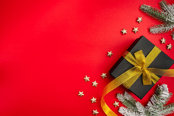 Elegant Christmas background with black gift box tied with gold ribbon, snowy pine branches, black baubles, and stars on red surface. Ideal for luxury holiday cards, banners, and festive promotions.