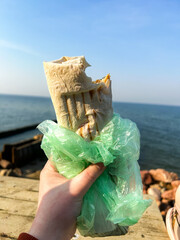 Woman holds Shawarma Wrap Kebab in hands in front of sea. Street food concept. To go food. Food on the go. Shawarma in lavash. Convenient food.