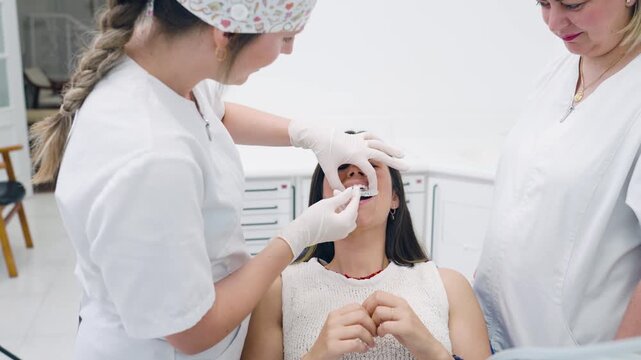Young female orthodontist, assisted by a colleague, fitting a clear aligner on a woman's teeth for a bite correction treatment