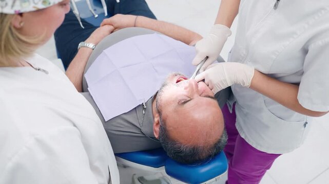 Professional dentist, with an assistant, performing a tooth extraction procedure on a mature male patient lying in a dental chair
