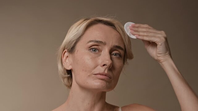 Static shot of blonde woman using cotton pad to apply toner on face while doing skincare routine
