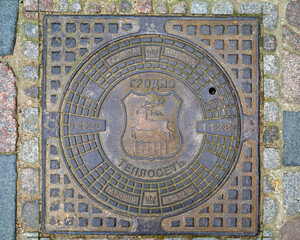 Round lid of a cast-iron manhole of a street sewer with the image of a deer and the inscription in Cyrillic Grodno and Latin letters on a paved street