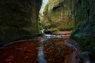 Scenic view of Finnich Glen in Scotland with moss-covered rock walls, flowing river and vibrant red sandstone. Atmospheric natural landscape and unique geological formation.