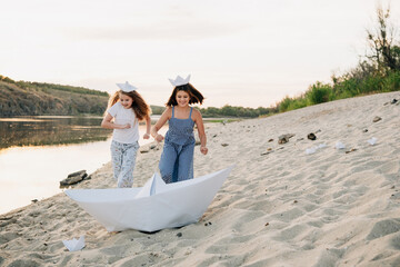 Two cheerful girls wearing paper hats and smiling, running barefoot on a sandy beach next to a large paper boat, playing by the river at sunset with small paper boats scattered around