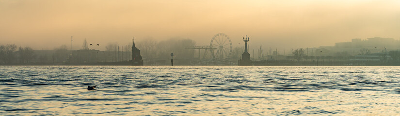 On a foggy winter day in pastel colors, seagulls fly along the waterfront with the port, the Imperia statue, and the Ferris wheel in the background. Konstanz, Baden-W&uuml;rttemberg, Germany.
