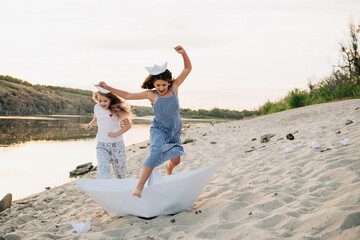 Two joyful girls running and jumping on a sandy beach next to a river, engaging in creative play with large and small paper boats, celebrating childhood and imagination during a golden hour sunset