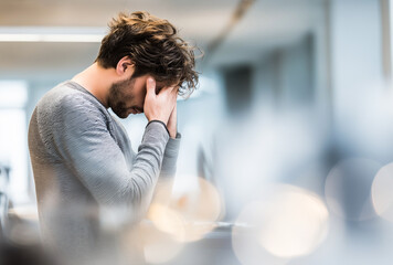Fototapeta premium Close-up of a distressed young man with brown hair holding his face in a blurred, bright office environment from a slightly low angle.