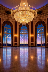 A luxurious ballroom, featuring a grand crystal chandelier and marble floor, has tall arched windows where light trails have drawn the number '2026' against a sunset sky. 
