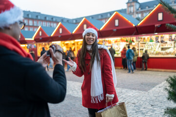 Man recording video of happy woman wearing santa hat at Christmas market, enjoying festive holiday season shopping
