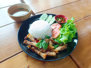  pork with basil topped with rice, vegetables, tomato slices, cucumber slices and dipping sauce in a red bowl, Placed on a black ceramic plate, and soup in a cup, on a wooden table background