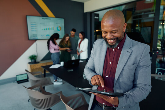 Happy black mature businessman using tablet with team discussing in background at workplace - Powered by Adobe