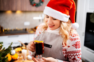 Woman smiling and holding hot mulled wine in christmas and new year kitchen with citrus and spices creating warm holiday atmosphere and cozy winter festive mood