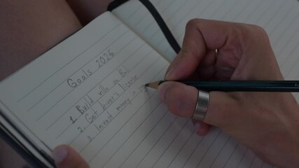 Close-up of a hand writing a 2026 personal goals list with a pencil in a lined notebook, focusing on detail and handwriting. 4K