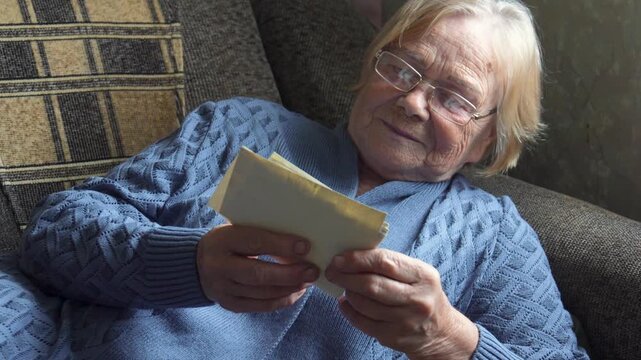 Old woman reading handwritten letter in cozy armchair wearing blue sweater and glasses. Soft window light gentle smile turning pages reflective mood. Vintage envelope paper, quiet domestic living room