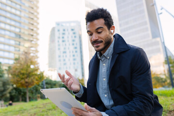 Young adult Latin American businessman talking while using tablet. Engaged professional communicating during outdoor work. Business and people concept.