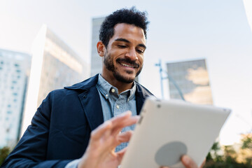 Young adult Latin American businessman using tablet in modern city area. Concentrated moment blending productivity and urban energy. Business and people concept.