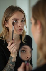 Young blonde woman applies eye makeup with brush. Looks into large mirror carefully. Small magnifying mirror helps see facial details clearly. Performs daily beauty routine focusing on pretty eyes.