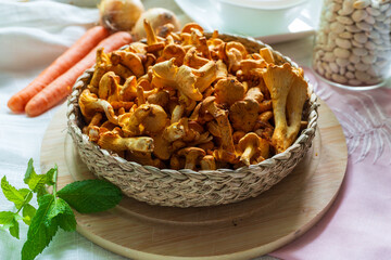 A woven basket overflowing with fresh chanterelle mushrooms is placed beside carrots, onions, beans, and mint leaves. A warm kitchen still life perfect for cooking, recipe development, and farm-to-tab