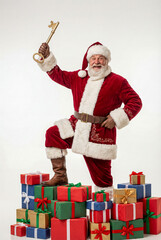 Santa Claus stands triumphantly on a large stack of wrapped, colorful presents, raising a giant golden key above his head against a stark white backdrop.