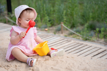 adorable toddler in pink outfit and sun hat plays on sandy boardwalk with red plastic shovel and yellow bucket, surrounded by lush green grass on a sunny day outdoors, playful, soft, individual