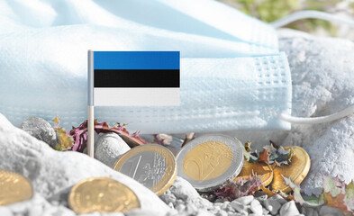 Estonian flag with euro currency coins and a medical mask on rocky ground.Eurozone healthcare costs,medical funding challenges and the financial weight of modern services connected to Estonia.