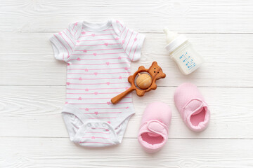 Baby bodysuits and eco friendly accessories - booties, wooden rattle toy and baby bottle with milk on white wooden background, top view