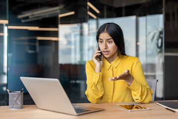 Worried young Indian woman sitting at desk in office and talking emotionally on phone