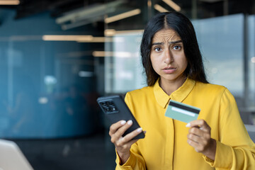 Close-up portrait of a young Indian upset woman in the office, holding a phone and credit card, looking at the camera