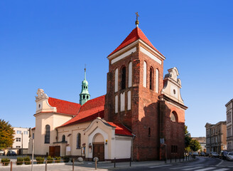 Fototapeta premium Front View of the Roman Catholic Church in Gniezno, Greater Poland Voivodeship with Red Clay Roof & Red Brick Tower Under Clear Blue Sky.