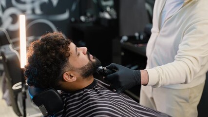 Professional barber trimming man's beard with electric shaver in barbershop