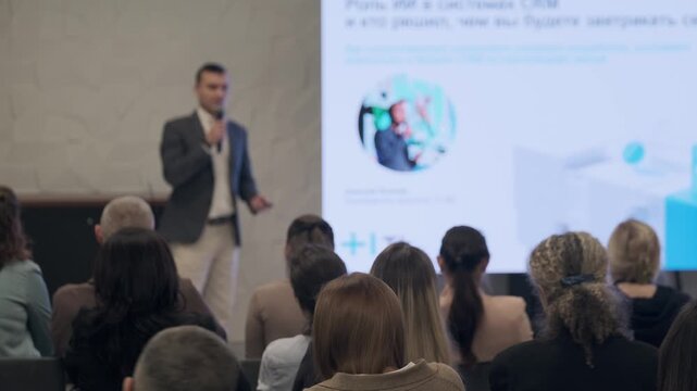 Presenter speaks to audience during conference. Attentive crowd sits facing stage while large screen shows graphics and text. Professional atmosphere emphasizes knowledge sharing collaboration 