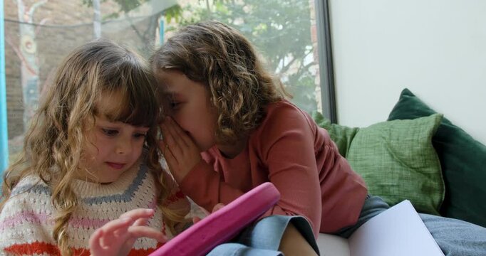 Girl whispering in Ear of Sister using Digital tablet in cozy corner