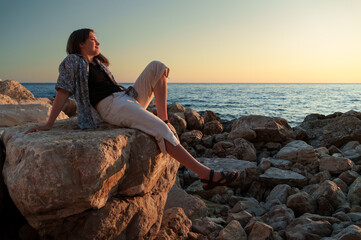 A girl poses and relaxes by the sea, sitting on rocks near the slope of a high cliff, panoramic view of the seashore and mountains at sunset