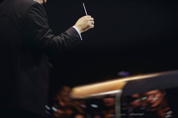 Male conductor behind the podium directing orchestra on stage with pupitre, musicians and choir,...